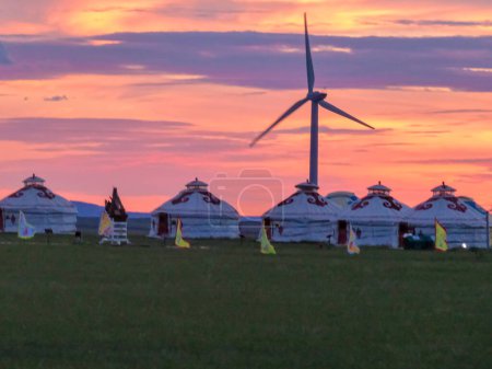 Foto de White traditional yurts located on a pasture in Xilinhot in Inner Mongolia  seen during the pink sunset. Endless grassland. Wind turbines in the back. Clean energy. Nomadic way of life. Natural wonder - Imagen libre de derechos