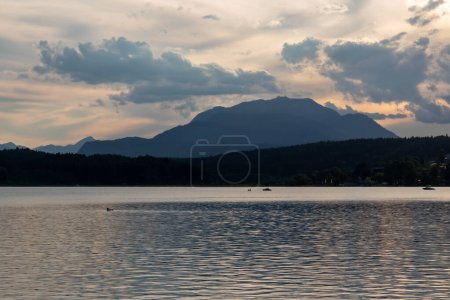 Photo for A panoramic view on the Lake Faak in Austria. The lake is surrounded by high Alpine peaks. The sun in slowly setting behind the mountains. Lots of clouds. Calm surface reflects the sunbeams. Serenity - Royalty Free Image