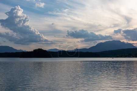 Photo for A panoramic view on the Lake Faak in Austria. The lake is surrounded by high Alpine peaks. The sun in slowly setting behind the mountains. Lots of clouds. Calm surface reflects the sunbeams. Serenity - Royalty Free Image