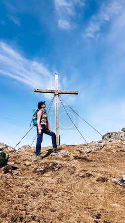 Foto de A woman with hiking backpack standing under a wooden cross on top of Sauofen in Austrian Alps. A vast, golden pasture around. Fall vibes. She is laughing, being happy. Reaching the top. Serenity - Imagen libre de derechos