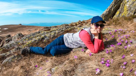 Foto de A woman lying on the golden pasture, surrounded by purple crocuses in the region of Sauofen in Austrian Alps. Spring on Alpine meadows. There is a sharp stony wall behind her. Smiling woman - Imagen libre de derechos