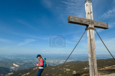 Foto de A woman with hiking backpack standing under a wooden cross on top of Sauofen in Austrian Alps. A vast, golden pasture around. Fall vibes. Mountain chains in the back. Panoramic view. Achievement - Imagen libre de derechos