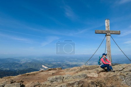 Foto de A woman with hiking backpack sitting under a wooden cross on top of Sauofen in Austrian Alps. A vast, golden pasture around. Fall vibes. Mountain chains in the back. Panoramic view. Achievement - Imagen libre de derechos