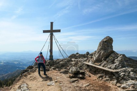 Foto de A woman with hiking backpack reaching the top of Sauofen in Austrian Alps. A wooden cross on top. Lots of stones around. Fall vibes. High mountain chains in the back. Serenity and achievement - Imagen libre de derechos