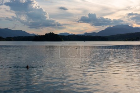 Photo for A woman swimming in the Lake Faak in Austria. The lake is surrounded by high Alpine peaks. The sun in slowly setting behind the mountains. Lots of clouds. Calm surface reflects the sunbeams. Happiness - Royalty Free Image