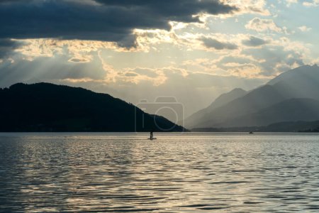 Photo for A man stand up paddling during the sunset on Millstaetter lake in Austria. The lake is surrounded by high Alps. Calm surface of the lake reflecting the sunbeams. The sun sets behind the mountains. - Royalty Free Image