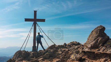 Foto de A man with hiking backpack standing on sharp rocks, under a wooden cross on top of Sauofen in Austrian Alps. A vast, golden pasture around. Fall vibes. Mountain chains in the back. Serenity - Imagen libre de derechos