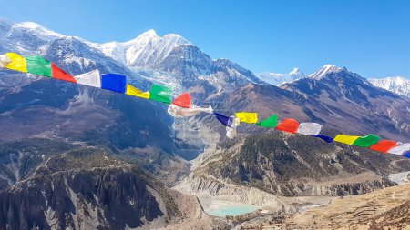 Foto de A panoramic view on Manang valley from Praken Gompa, Nepal. Prayer flags waving above the snow capped peaks of Annapurna Chain. There is a small lake in the valley. Harsh landscape. Serenity - Imagen libre de derechos