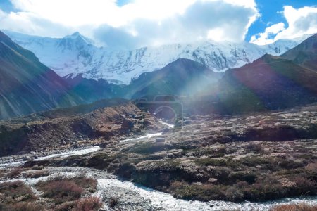 Téléchargez les photos : Vue panoramique sur une vallée le long du circuit de l'Annapurna au Népal, avec un petit torrent qui coule au milieu. À l'arrière, il y a de hauts sommets himalayens enneigés. Les pentes sont envahies par de petits buissons - en image libre de droit