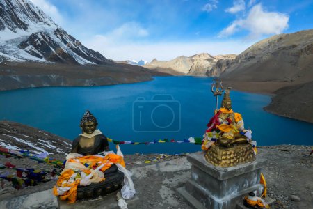 Foto de Dos estatuas de Buda en el lago Tilicho, cubiertas con banderas de oración. Superficie azul y tranquila del lago, montañas cubiertas de sombra, luz solar en la parte posterior. Annapurna Circuit Trek, Nepal. La espiritualidad - Imagen libre de derechos