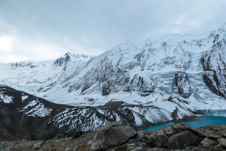 Téléchargez les photos : Stormy weather in snow capped Himalayan peaks along Annapurna Circuit in Nepal. Barren and sharp slopes. Mountains are partially shrouded with clouds. There is a turquoise Tilicho Lake on the side. - en image libre de droit