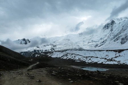Téléchargez les photos : Stormy weather in snow capped Himalayan peaks along Annapurna Circuit in Nepal. Barren and sharp slopes. Mountains are partially shrouded with clouds. There is small pathway. Tilicho Lake on the side - en image libre de droit