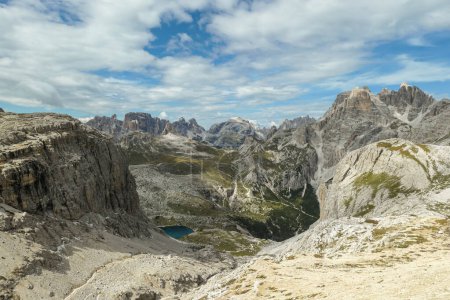 Foto de Una vista panorámica de los Dolomitas en Italia. Hay fuertes y empinadas laderas de montaña alrededor. En el fondo de un pequeño valle hay un pequeño lago azul marino. El cielo está lleno de nubes suaves. Paisaje crudo - Imagen libre de derechos