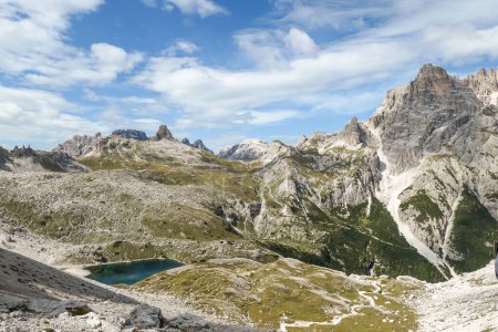 Foto de Una vista panorámica de los Dolomitas en Italia. Hay fuertes y empinadas laderas de montaña alrededor. En el fondo de un pequeño valle hay un pequeño lago azul marino. El cielo está lleno de nubes suaves. Paisaje crudo - Imagen libre de derechos