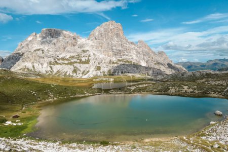 Foto de Un pequeño lago azul marino en el fondo del valle en los Alpes italianos. El lago está rodeado de picos altos y empinados. Las laderas son de un verde exuberante. El cielo está lleno de nubes suaves. Paisaje crudo. Remedio - Imagen libre de derechos