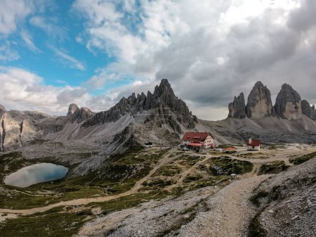 Téléchargez les photos : Une capture du chalet Tre Cime di Lavaredo (Drei Zinnenhuette) dans les Dolomites italiennes avec un lac turquoise derrière. Le chalet a des éléments décoratifs rouges. Il y a de hauts sommets alpins autour. Abri - en image libre de droit