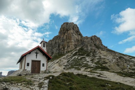 Foto de Una captura de una pequeña capilla, con tejado rojo y pequeño campanario en los Dolomitas italianos. Hay altos picos alpinos alrededor. Un poco nublado. Espiritualidad y meditación. Refugio - Imagen libre de derechos