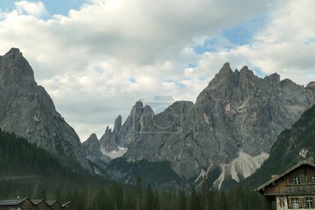 Foto de Una vista de cerca de los picos altos y agudos en los Dolomitas italianos. Las laderas tienen muchos derrumbes y pierden rocas. Partes inferiores cubiertas de bosque denso. Unos cuantos tejados al frente. Pocas nubes arriba - Imagen libre de derechos