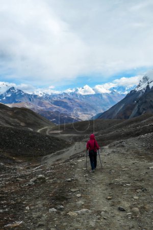 Téléchargez les photos : A woman in pink jacket walking on a pathway leading to Tilicho Lake, Nepal. Stormy weather in snow capped Himalayan peaks along Annapurna Circuit. Barren and sharp slopes. Coldness - en image libre de droit