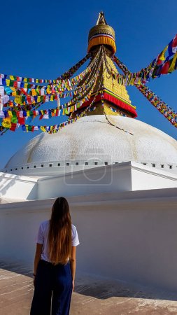 Foto de A girl with long hair standing in front of boudhanath temple in Kathmandu, Nepal. The temple is painted white. There are thousands of prayer flags attached to the top of the temple. Spirituality. - Imagen libre de derechos