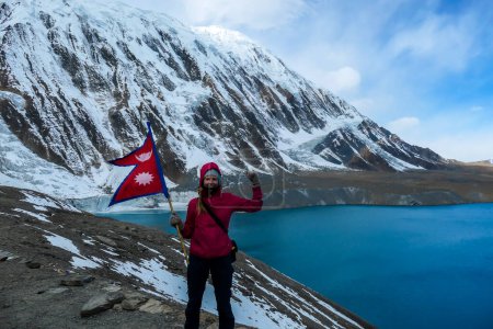 Téléchargez les photos : A woman holding Nepalese flag while standing at the side of turquoise colored Tilicho lake in Himalayas, Manang region in Nepal. The world's highest altitude lake. Snow capped mountains around. - en image libre de droit