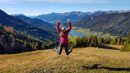 Téléchargez les photos : A woman in pink jacket jumping with joy while hiking the view on a distant Weissensee lake. The grass is golden, trees also change colors to yellow and orange. High Alps around. Autumn vibes - en image libre de droit