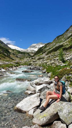 Photo for Woman wearing a backpack enjoying a cascading waterfall in the Alps in Ankogel group in Austria. The rushing torrent is coming from a glacier water. High mountains around, overgrown with small bushes - Royalty Free Image