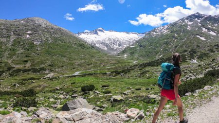 Photo for A woman wearing shorts and hiking backpack hiking in Austrian Alps, along a gravelled road. High mountains around. There is a glacier in the back. Adventure and discovery. Blossoming Alpine meadow - Royalty Free Image