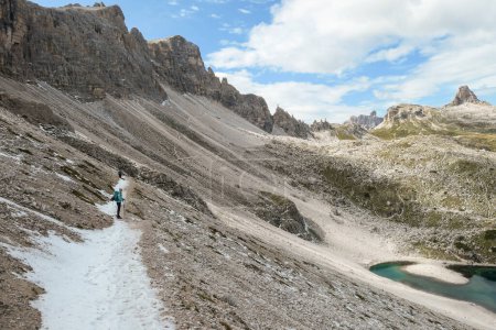 Foto de Una mujer con mochila y palos de senderismo en un camino estrecho y helado en Dolomitas italianas. Hay montañas afiladas y empinadas. En el fondo de un pequeño valle hay un pequeño lago azul marino. Paisaje crudo - Imagen libre de derechos