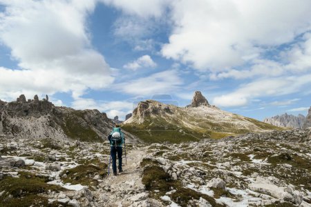 Photo for A woman with backpack and sticks hiking on a narrow path in Italian Dolomites. There are sharp and steep mountains. At the bottom of a small valley there is a small, navy blue lake. Raw landscape. - Royalty Free Image