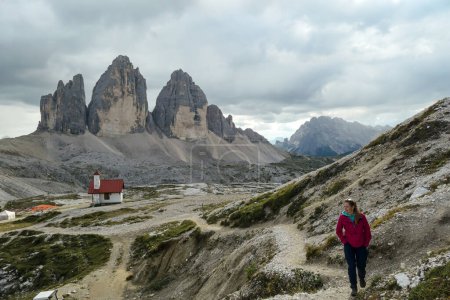 Photo for A woman in pink jacket hiking with a small chapel, with red rooftop behind her and the view on the Tre Cime (Drei Zinnen) in Italian Dolomites. There are high Alpine peaks around. A bit of overcast. - Royalty Free Image