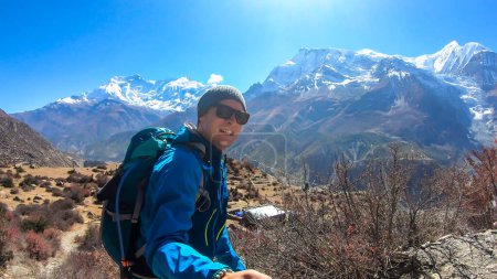 Foto de A man hiking and taking a selfie with snow caped Annapurna chain in the back, Annapurna Circuit Trek, Himalayas, Nepal. High mountains around. The man is admiring the landscape. Serenity and calmness. - Imagen libre de derechos