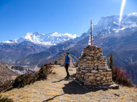 Ein junger Mann steht neben einer Reihe kleiner steinerner Stupas mit der Annapurna-Kette als Hintergrund, Himalaya, Nepal. Hohe schneebedeckte Berge. Karges und trockenes Land. Eine Gebetsfahne daneben.