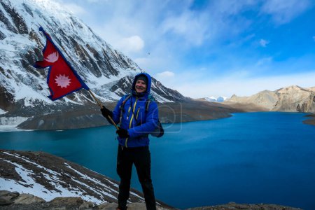 Téléchargez les photos : Un homme tenant le drapeau népalais debout au bord du lac Tilicho de couleur turquoise dans l'Himalaya, région de Manang au Népal. Le lac d'altitude le plus élevé du monde. Montagnes enneigées autour. - en image libre de droit