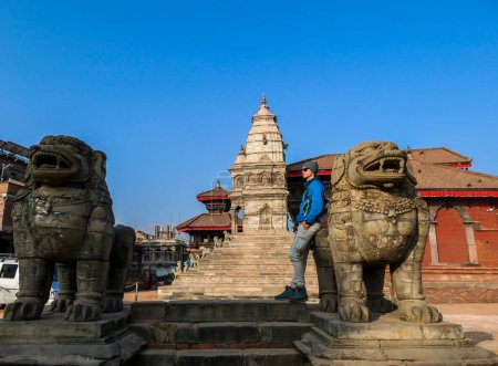 Téléchargez les photos : A man leaning against a guardian lion with the view on Stupa in t in Bhaktapur, Nepal.There are two guardian lions. Man enjoys early morning on a quiet main square in ancient city. Serenity - en image libre de droit