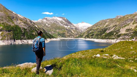 Photo for Man standing at the side of a lake at Koelnbreinsperre dam in Austria. The lake has navy blue color. High Alps around. There is a glacier in the back. The meadows blossoming with wild flowers. Calmness - Royalty Free Image
