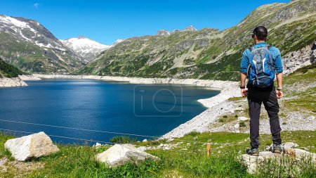 Photo for Man standing at the side of a lake at Koelnbreinsperre dam in Austria. The lake has navy blue color. High Alps around. There is a glacier in the back. The meadows blossoming with wild flowers. Calmness - Royalty Free Image