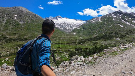 Photo for Man with hiking backpack taking a selfie while hiking in Austrian Alps, along a gravelled road. High mountains around. There is a glacier in the back. Adventure and discovery. Clear and bright day - Royalty Free Image