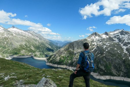 Photo for Man with hiking backpack admiring a view on an artificial lake at a dam in Austria from above. The lake has navy blue color. High Alps around. There is a glacier in the back. Adventure and discovery - Royalty Free Image