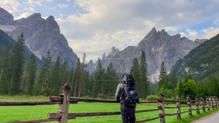Foto de Un hombre con una gran mochila de senderismo disfrutando de la vista lejana de los Dolomitas italianos. Hay un exuberante prado verde con una valla de madera a su alrededor. Niebla matutina en el valle. Bosque grueso en las laderas - Imagen libre de derechos