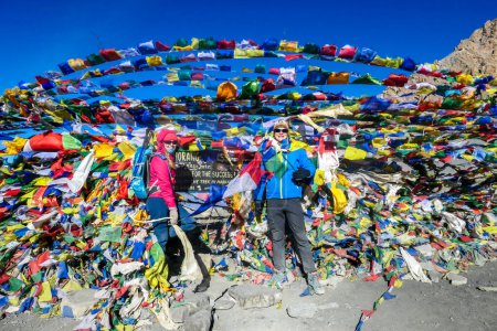 A couple standing between prayer flags at the top of Thorung La Pass, Annapurna Circuit Trek, Nepal. Memorial board. Colorful prayer flags attached to the stone wall, blown by the wind. Clear sky.