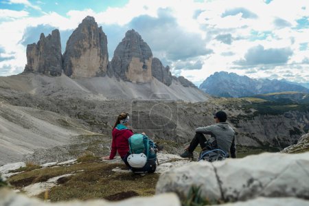 Téléchargez les photos : A couple in hiking outfit, sitting on the ground and enjoying the view on the famous Tre Cime di Lavaredo (Drei Zinnen) in Italian Dolomites. Desolated and raw landscape, full of lose stones. Overcast - en image libre de droit