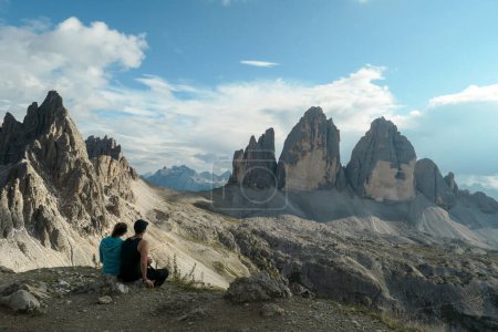 Téléchargez les photos : A couple in hiking outfit, sitting on the ground and enjoying the view on the famous Tre Cime di Lavaredo (Drei Zinnen) in Italian Dolomites. Desolated, raw landscape, full of lose stones. Sunny day - en image libre de droit