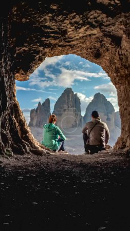 Foto de Una pareja sentada frente a una cueva, con la vista sobre el Tre Cime (Drei Zinnen) en los Dolomitas italianos. Están disfrutando del paisaje tranquilo. Picos de montaña empinados y agudos. Serenidad y meditación - Imagen libre de derechos