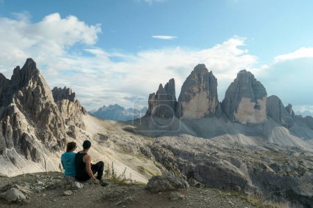 Téléchargez les photos : A couple in hiking outfit, sitting on the ground and enjoying the view on the famous Tre Cime di Lavaredo (Drei Zinnen) in Italian Dolomites. Desolated, raw landscape, full of lose stones. Sunny day - en image libre de droit