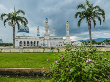 Foto de Masjid Bandaraya en Kota Kinabalu, Sabah, Malasia. Césped verde exuberante frente a la mezquita. Dos palmeras a los lados del templo. Un arbusto de flores en el frente. Enorme nublado, la tormenta se acerca. - Imagen libre de derechos