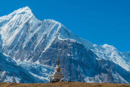 Foto de Una estupa con Annapurna Chain como telón de fondo, Annapurna Circuit Trek, Himalaya, Nepal. Montañas altas cubiertas de nieve. La tierra frente a la estupa es estéril y seca. Bandera de oración junto a ella. - Imagen libre de derechos