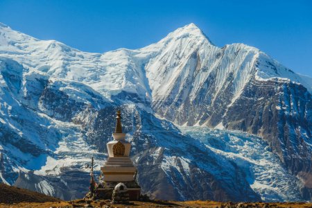 Una estupa con Annapurna Chain como telón de fondo, Annapurna Circuit Trek, Himalaya, Nepal. Montañas altas cubiertas de nieve. La tierra frente a la estupa es estéril y seca. Bandera de oración junto a ella.
