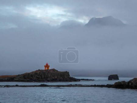 Foto de A shore of a fjord. At one of the headlands there is a small orange lighthouse. In the back higher mountains hiding in the clouds. Calm water surrounding the headlands. - Imagen libre de derechos