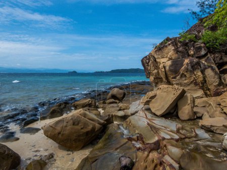 Foto de Una hermosa formación rocosa junto a la playa en Malasia. Las piedras son lavadas por las olas, se ven muy resbaladizas y peligrosas. Encima de las rocas hay algunos arbustos. Suaves olas corriendo a la orilla - Imagen libre de derechos